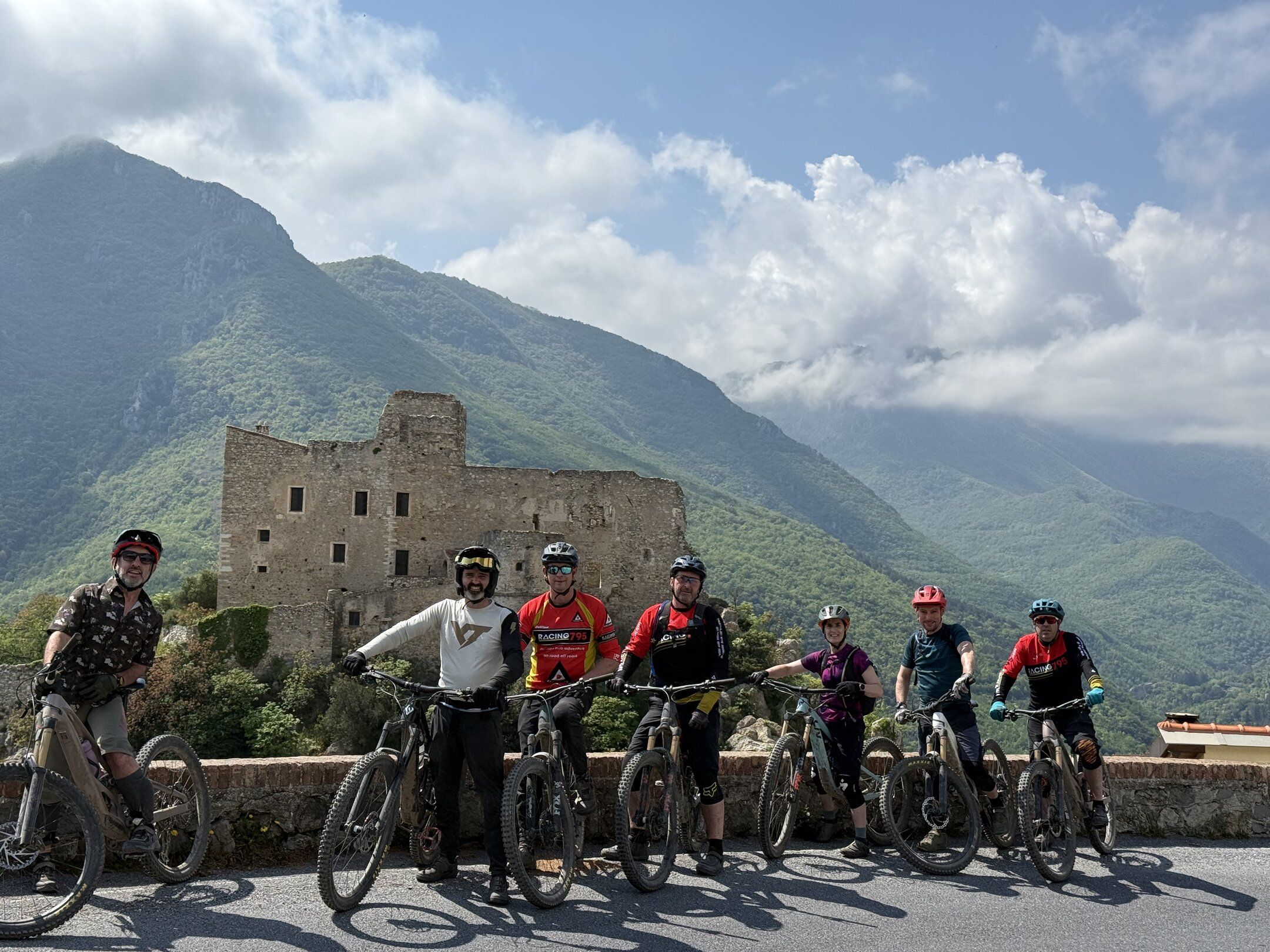 A group of riders with their mountain bikes in front of a ruined castle, mountains behind